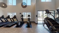 This contemporary fitness center is designed to support a comprehensive workout experience within a spacious, single-floor layout. The facility is immediately distinguished by its dramatically high ceilings, which create an open, airy atmosphere throughout the space. Large windows line the walls, flooding the interior with natural light and offering views of the surrounding outdoor courtyard and community areas. Stylish woven globe pendant lights serve as decorative focal points, complementing the modern aesthetic of the clean white walls and neutral-toned flooring.

The cardio zone is generously equipped with multiple treadmills, elliptical trainers, and StairMaster steppers, all arranged to take advantage of the window views. A dedicated strength training area features cable machines, adjustable weight benches, dumbbells, and kettlebells neatly organized on racks, along with wall-mounted mirrors to assist with form and technique. A flat-screen television is mounted on the wall for entertainment during workouts.

A separate, open studio area with smooth flooring and a ceiling fan provides space for stretching, yoga, or group fitness activities, with floor-to-ceiling glass doors that open directly to the outdoor patio. Additional amenities include a water fountain and a branded entrance mat. The facility is thoughtfully designed to be both functional and visually inviting.