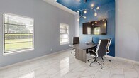 Located at 3205 E Olive Rd in Pensacola, Florida, this thoughtfully designed office space makes efficient use of its compact footprint to deliver a polished and functional work environment. The single-room layout is divided into two distinct work zones, each with its own character and purpose.

The primary workstation area features a sleek, angular wood-toned desk surrounded by ergonomic high-back black rolling chairs, accommodating small team collaboration or focused individual work. A large flat-screen display is mounted on a striking deep blue accent wall, complemented by a bold circular architectural feature with a warm wood-inlay surround — a standout design element that adds visual depth and sophistication.

An overhead branching chandelier with globe bulbs provides warm, ambient lighting above this zone, while the vaulted ceiling painted in a rich blue tone enhances the sense of height and drama. Adjacent to this, a shared workstation along the wall offers additional seating and desktop computer access, illuminated by sleek modern pendant lights.

The flooring throughout is polished white marble-look tile, lending the space a clean, contemporary aesthetic. Light gray walls with white trim keep the overall palette fresh and professional. Natural light filters in through windows fitted with white blinds, and a glass-panel exterior door adds connectivity to the outdoors. Three decorative circular gold-framed mirrors accent one wall, adding an artistic touch to the workspace.