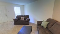 This single-floor space offers a comfortable and functional layout designed for everyday living. The living area is the focal point of the space, featuring soft, neutral-toned carpeting that adds warmth underfoot and contributes to an inviting atmosphere. The walls are finished in a clean, light neutral tone, providing a versatile backdrop that enhances the sense of openness throughout the room. A window dressed with vertical blinds allows natural light to filter into the space, brightening the interior while offering privacy and light control. The textured ceiling adds a subtle architectural detail to the room. The living area provides ample room for seating and a central coffee table, making it well-suited for relaxing or entertaining. The overall ambiance of the space is understated and practical, with a calm, neutral palette that lends itself to a variety of interior styles. The layout and design reflect a straightforward, livable space that prioritizes comfort and functionality in a compact, single-level footprint.
