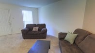 This single-floor space offers a comfortable and functional layout designed for everyday living. The living area is the focal point of the space, featuring soft, neutral-toned carpeting that adds warmth underfoot and contributes to an inviting atmosphere. The walls are finished in a clean, light neutral tone, providing a versatile backdrop that enhances the sense of openness throughout the room. A window dressed with vertical blinds allows natural light to filter into the space, brightening the interior while offering privacy and light control. The textured ceiling adds a subtle architectural detail to the room. The living area provides ample room for seating and a central coffee table, making it well-suited for relaxing or entertaining. The overall ambiance of the space is understated and practical, with a calm, neutral palette that lends itself to a variety of interior styles. The layout and design reflect a straightforward, livable space that prioritizes comfort and functionality in a compact, single-level footprint.