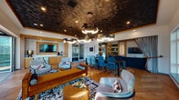Located at 3883 S Moody Ave in Portland, Oregon, this sophisticated single-floor communal space delivers a striking blend of contemporary design and refined comfort. The open-plan layout seamlessly connects multiple functional zones, creating an inviting atmosphere suited for both socializing and relaxation.

The space is immediately distinguished by its dramatic textured ceiling, finished in a dark geometric pattern that adds depth and visual intrigue throughout the main area. A sculptural cluster of modern ring-style pendant lights anchors the room, casting warm, layered illumination across the herringbone-patterned hardwood floors below.

The dining area features a substantial dark wood table surrounded by teal upholstered chairs, providing ample seating for gatherings. Adjacent to it, a lounge zone offers a curated arrangement of light gray armchairs, a rich caramel-toned sofa, and a bold geometric area rug, all centered around a round coffee table. Built-in shelving with teal-painted interiors lines one wall, adding both storage and a vibrant design accent.

A secondary lounge area is defined by a deep navy sectional sofa and a wall-mounted television set within warm wood-framed paneling. Large floor-to-ceiling windows throughout the space flood the interior with natural light and frame panoramic views of the surrounding Portland waterfront and cityscape. Drapery panels provide an elegant finishing touch, offering privacy without sacrificing the expansive, airy feel of the room.