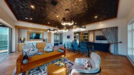 Located at 3883 S Moody Ave in Portland, Oregon, this sophisticated single-floor communal space delivers a striking blend of contemporary design and refined comfort. The open-plan layout seamlessly connects multiple functional zones, creating an inviting atmosphere suited for both socializing and relaxation.

The space is immediately distinguished by its dramatic textured ceiling, finished in a dark geometric pattern that adds depth and visual intrigue throughout the main area. A sculptural cluster of modern ring-style pendant lights anchors the room, casting warm, layered illumination across the herringbone-patterned hardwood floors below.

The dining area features a substantial dark wood table surrounded by teal upholstered chairs, providing ample seating for gatherings. Adjacent to it, a lounge zone offers a curated arrangement of light gray armchairs, a rich caramel-toned sofa, and a bold geometric area rug, all centered around a round coffee table. Built-in shelving with teal-painted interiors lines one wall, adding both storage and a vibrant design accent.

A secondary lounge area is defined by a deep navy sectional sofa and a wall-mounted television set within warm wood-framed paneling. Large floor-to-ceiling windows throughout the space flood the interior with natural light and frame panoramic views of the surrounding Portland waterfront and cityscape. Drapery panels provide an elegant finishing touch, offering privacy without sacrificing the expansive, airy feel of the room.
