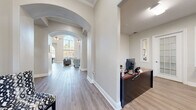 This single-floor facility offers a thoughtfully designed layout spanning a generous footprint, blending professional functionality with inviting architectural character. The entrance area greets visitors with elegant arched doorways, crown molding, and light wood-tone flooring that flows seamlessly throughout the space, creating a cohesive and polished aesthetic.

The centerpiece of the facility is a dramatic great room distinguished by soaring vaulted ceilings, a striking floor-to-ceiling arched window arrangement that floods the space with natural light, and a classic fireplace. A ceiling fan and recessed lighting complement the room's airy, open atmosphere.

Two dedicated office spaces provide practical work environments. The primary office features a reception-style desk setup, direct exterior access via a glass-paneled door, and wall-mounted décor. The secondary office offers a built-in workstation along one wall, ample natural light from multiple windows, and track lighting overhead — well-suited for collaborative or individual work.

The exercise room is equipped with multiple treadmills, a multi-station cable machine, free weights, and wall-to-wall mirrors, all set on durable flooring with bright overhead lighting and windows providing natural ventilation.

Five hallways connect the various areas of the facility, with welcoming nooks featuring accent furniture, art, and natural light, contributing to a warm and professional overall ambiance throughout.