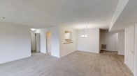 This single-floor space features an open-concept layout that connects the living, dining, and kitchen areas in a cohesive and functional arrangement. The living and dining areas are finished with neutral-toned wall-to-wall carpeting, contributing to a warm and understated atmosphere throughout.

A defining architectural feature is the partial dividing wall with a pass-through window that connects the kitchen to the main living area. This design element allows for easy interaction between spaces while maintaining a degree of separation. The kitchen, visible through the pass-through, features white cabinetry and appears to include standard appliances.

The dining area is highlighted by a modest chandelier, providing a focal point and adding a touch of elegance to the otherwise simple aesthetic. The walls are painted in a clean, bright white throughout, enhancing the sense of openness and light.

A hallway visible on the left side of the space leads to additional rooms, with a door and overhead lighting indicating access to private areas of the unit. The textured ceiling is consistent throughout the main living areas. A floor-level vent indicates a central HVAC system. Overall, the space presents a clean, move-in-ready environment with a simple and functional design.