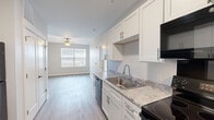Located at 419 Donald Driver Way in Green Bay, Wisconsin, this 1-bedroom, 1-bathroom unit offers a thoughtfully organized single-floor layout with modern finishes throughout. The open-concept kitchen and living area creates a cohesive, functional space anchored by light wood-look flooring and a ceiling fan. The kitchen is well-appointed with white shaker-style cabinetry, granite-look countertops, a stainless steel sink, and black appliances including a refrigerator, electric range, built-in microwave, and dishwasher. Additional storage is provided by a full-height pantry cabinet near the entryway. The living area receives natural light through a large window with horizontal blinds, contributing to a bright and airy atmosphere. The bedroom features soft carpeting and a door leading to a private outdoor entry, allowing for additional natural light. A closet provides dedicated storage within the bedroom. The bathroom is cleanly finished with wood-look flooring, a white vanity with a solid surface countertop, a bathtub with grab bar, and a stacked in-unit washer and dryer, adding everyday convenience. Neutral wall tones and clean white trim carry throughout the space, lending a cohesive and move-in-ready aesthetic.