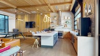 Located at 2025 50th Avenue in Greeley, Colorado, this thoughtfully designed single-floor space seamlessly blends the kitchen and living areas into one cohesive, open-concept layout. The interior is immediately distinguished by its striking wood-planked ceiling with exposed beams, which adds warmth and architectural character throughout. Wide-plank hardwood flooring ties the entire space together, providing a natural, grounded foundation underfoot.

The kitchen is a functional centerpiece, featuring a generous waterfall-edge island with seating for multiple guests, warm wood cabinetry, white quartz countertops, a subway tile backsplash, open floating shelves, a stainless steel range, and a sleek wall-mounted range hood. Large garage-style windows flood the kitchen with natural light and offer views of the surrounding neighborhood.

The living area transitions effortlessly from the kitchen, highlighted by a dramatic floor-to-ceiling stacked-stone fireplace that serves as a bold focal point. Vertical wood slat wall paneling frames a mounted flat-screen television, flanked by contemporary lounge seating. A sculptural neon light fixture adds a playful, modern touch. Steel-framed glass doors connect to an adjacent room, enhancing the sense of openness and flow throughout this well-appointed, design-forward space.