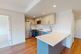 This modern 1-bedroom, 1-bathroom residence in Cambridge, MA offers a thoughtfully designed layout across a single floor. The open-concept kitchen and living area serves as the heart of the home, featuring warm hardwood flooring, recessed lighting, and a peninsula with a white quartz countertop that provides additional prep space and casual seating. The kitchen is well-appointed with two-tone cabinetry — white upper cabinets and slate-blue lower cabinets — complemented by stainless steel appliances including a refrigerator, range, dishwasher, and microwave. A bright hallway connects the main living area to the private bedroom, which is carpeted and bathed in natural light through large floor-to-ceiling windows overlooking the Cambridge cityscape. The bedroom also benefits from two closets for ample storage. The full bathroom includes a tub/shower combo with white tile surround, hardwood flooring, a white vanity with an illuminated medicine cabinet mirror, and clean, modern finishes throughout. A dedicated in-unit laundry area adds everyday convenience. The space maintains a crisp, contemporary aesthetic with white walls and an efficient, well-organized floor plan.