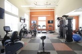This fitness center is designed to accommodate a comprehensive range of workout routines within a single, open floor plan. The space features soaring ceilings that create an airy, expansive feel, complemented by large multi-pane windows along multiple walls that allow abundant natural light to fill the room. Bold burnt-orange accent walls provide an energizing visual contrast against the neutral beige tones throughout.

The flooring consists of a distinctive checkered pattern of multi-toned interlocking tiles in orange, tan, brown, and gray, lending the space a dynamic aesthetic while providing a functional workout surface. Cardio equipment includes multiple treadmills and elliptical cross-trainers by Cybex, positioned to face wall-mounted flat-screen televisions for entertainment during workouts. A dedicated free weights area features a full dumbbell rack with an adjacent weight bench, while a multi-functional cable and strength training machine anchors the center of the room. An additional alcove houses medicine balls, stability balls, and supplementary training accessories. A wall of mirrors expands the perceived space and assists with form checks. Overhead linear lighting ensures consistent illumination throughout.
