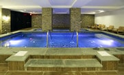 Located at 75 Sterling Blvd in Englewood, NJ, this single-floor indoor aquatic and wellness facility offers a thoughtfully designed environment centered around relaxation and therapeutic care. The space is anchored by a generously sized indoor pool with clear, illuminated water, complemented by a separate hydrotherapy spa area tucked within a warmly lit alcove framed by stacked stone accent columns. The architecture blends clean white walls with rich wood-look tile flooring and natural stone wall cladding, creating a serene and upscale ambiance throughout. Recessed lighting and elegant pendant fixtures provide warm, even illumination across the space. Along one side of the pool, a row of wooden lounge chairs and side tables invites relaxation before or after a swim. The facility is fully equipped with an ADA-compliant pool lift, ensuring accessibility for all guests. Individual rinse shower stations are conveniently positioned along the poolside wall, offering functional convenience without disrupting the flow of the space. Glass-panel partitions and dark wood cabinetry add refined architectural detail near the entrance area. The overall design strikes a balance between clinical functionality and spa-like comfort, making this a well-appointed destination for aquatic therapy, wellness, and leisure.