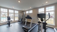 Located at Solomon Square in Delaware, Ohio, this single-floor community amenity space is designed to support an active lifestyle. The fitness center serves as the heart of the facility, offering a spacious, well-lit workout area with durable rubber flooring suited for high-impact activity. Large multi-pane windows line the walls, flooding the room with abundant natural light and providing views of the surrounding community. The gym is outfitted with a full array of equipment, including treadmills, elliptical machines, a multi-station cable machine, adjustable weight benches, and free weights, accommodating a wide range of fitness routines. A wall-mounted flat-screen television adds convenience for workout entertainment. Recessed lighting throughout ensures the space remains bright and functional at any hour. Elegant French doors with frosted glass panes separate the fitness area from an adjacent corridor, lending a polished architectural touch. A dedicated pet grooming room rounds out the amenity offerings, featuring a stainless steel grooming tub with a handheld sprayer and built-in open shelving for storing supplies. Light tile flooring and neutral wall tones keep the grooming space clean and practical. Together, these amenities reflect a thoughtfully designed community hub built for everyday comfort and convenience.