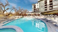 Located at Highline Pointe in Denver, Colorado, this resort-style outdoor amenity area offers a comprehensive recreational experience centered around a generously sized swimming pool with clear blue water. The pool deck is surrounded by an expansive concrete surround furnished with modern chaise lounges, side tables, and yellow-accented umbrellas that provide shade throughout the day. A dedicated hot tub is positioned adjacent to the main pool, offering a relaxing retreat year-round. The outdoor space also features thoughtfully designed cabana structures with warm wood slatted panels, angular steel frames, and string lighting overhead, creating intimate seating areas outfitted with contemporary white lounge furniture and bold yellow cushions. Raised concrete planters with native landscaping add texture and greenery throughout the space, while ornamental trees and perennial plantings provide natural screening and seasonal color. Outdoor dining tables and chairs are scattered across the deck, complementing the lounge areas for casual gathering. The entire amenity area is enclosed by black iron fencing and benefits from abundant natural light and sweeping views of the surrounding Denver skyline and mature tree canopy. The multi-story Highline Pointe building serves as an impressive architectural backdrop, completing this well-appointed outdoor retreat.