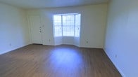 This single-floor space centers around a standout curved bay window that serves as the focal point of the main living area. The multi-paned window spans a generous portion of the exterior wall, allowing abundant natural light to pour into the room and offering outward views of surrounding greenery. The curved architecture of the bay adds visual interest and a sense of depth to the otherwise clean, understated layout.

The living area features smooth wood-look plank flooring in a warm, medium-toned finish that extends throughout the space, providing a cohesive and low-maintenance surface underfoot. The walls are finished in a crisp, neutral white, contributing to a bright and open atmosphere. A textured ceiling adds a subtle traditional detail overhead.

A white paneled door is positioned to the left of the bay window, providing access to an adjacent area or entry point. White baseboards frame the perimeter of the room, tying together the clean, uniform aesthetic. Overall, the space presents a functional and well-lit interior with simple, timeless finishes suited to a variety of furnishing styles.