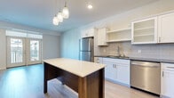 This single-story, one-bedroom, one-bathroom residence in Eagan, Minnesota, offers a thoughtfully designed layout with contemporary finishes throughout. Light-toned hardwood-style flooring runs continuously through the main living areas, creating a cohesive and clean aesthetic.

The kitchen is a standout feature, equipped with white shaker-style cabinetry, stainless steel appliances including a refrigerator, range, microwave, and dishwasher, complemented by a tile backsplash and dark countertops. A central island with a contrasting dark wood base and light countertop provides additional workspace and serves as a natural divider between the kitchen and living area. Pendant lighting overhead adds a modern touch. A built-in desk area and tall storage cabinet near the entry offer practical organizational solutions.

The living area is bright and open, benefiting from natural light streaming through large windows and a glass door leading to a private balcony with outdoor views. The bathroom features a framed mirror, vanity with cabinetry, and wood-look flooring. A dedicated in-unit laundry space houses a full-sized washer and dryer with overhead shelving for added storage convenience.
