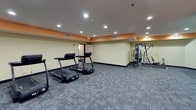 This fitness center is designed to accommodate a well-rounded workout experience within a single-level layout. The space is finished with durable, patterned carpet tiles in a gray tone throughout, providing a practical and clean foundation underfoot. Recessed lighting illuminates the entire area evenly, and a distinctive orange accent band runs along the upper portion of the light yellow walls, giving the space a vibrant, energetic character.

The cardio zone is equipped with multiple treadmills and two elliptical trainers, offering ample options for aerobic exercise. A multi-station cable strength machine and additional weight equipment occupy a dedicated area along one wall, catering to resistance and strength training needs. A workout chart is posted nearby for reference.

A separate, open area within the space serves as a stretching and flexibility zone, furnished with exercise mats laid on the floor, resistance hoops, a stability ball, and a low bench for accessories. This section features a curved wall detail that adds architectural interest.

Ventilation is supported by ceiling-mounted HVAC vents throughout, and an emergency exit door is accessible from the main workout area, ensuring safety and convenience for all users.