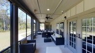 This single-story property features a generously proportioned screened porch that serves as a standout outdoor living area. The porch is enclosed with full-length screen panels framed in dark trim, allowing unobstructed views of the surrounding landscape, including open lawn and mature trees. The structure is supported by board-and-batten siding in a neutral tone, lending a clean, modern farmhouse aesthetic.

The ceiling is finished with a flat panel design and fitted with recessed lighting and two ceiling fans, ensuring comfort and ventilation throughout the warmer months. A security camera is also mounted, adding a practical element to the space.

The porch is furnished with dark wicker sofas and chairs accented by white and lime green cushions, arranged around a wicker coffee table with a decorative lantern centerpiece. The concrete flooring provides a durable, low-maintenance surface that complements the overall design.

Access to the interior of the building is provided through multiple French-style doors along the interior wall, maintaining a seamless connection between indoor and outdoor spaces. The abundance of natural light and open sightlines create a bright, airy ambiance throughout.