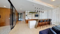 This club room is a thoughtfully designed communal space that blends modern aesthetics with warm, inviting materials. The layout encompasses several distinct functional areas, each with its own character while maintaining a cohesive design language throughout.

The main lounge area is bright and airy, featuring large glass doors that flood the space with natural light and provide views to an outdoor terrace. Comfortable blue velvet sofas are arranged around a bold yellow area rug, accompanied by a wall-mounted television and a cluster of smoked-glass pendant lights overhead, creating an ideal space for relaxed social gatherings.

Adjacent to the lounge is a stylish bar and counter area, anchored by a sleek white island and flanked by warm walnut cabinetry with gold hardware. A gold-framed open shelving unit adorned with trailing greenery and decorative accents serves as a striking focal point, while pendant lights in varying styles hang above bar-height seating.

A private meeting room offers a more intimate setting, centered around a polished black dining table with upholstered chairs. A linear chandelier of globe pendants illuminates the space, complemented by bold abstract artwork on the wall.

A dedicated kitchenette with walnut cabinetry and a microwave rounds out the amenities. Large-format tile flooring and sheer drapery unify the entire space with an understated elegance.