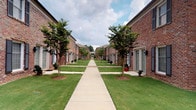 This courtyard space is defined by its classic brick construction and open-air orientation, offering a functional and visually connected outdoor area. The covered portion features exposed brick walls displaying a rich, variegated pattern of red and earth-toned bricks, lending the space a traditional, established character. The concrete floor extends throughout, providing a durable, level surface suitable for a variety of uses.

The covered area opens directly onto a well-maintained central walkway that serves as the spine of the surrounding community. Flanking this pathway are two rows of brick buildings of consistent architectural style, each fronted by neatly trimmed lawns and young crape myrtle trees planted at regular intervals. The trees provide seasonal interest and a sense of scale along the corridor, while gray entry doors and shuttered windows contribute a uniform, orderly aesthetic.

From within the covered space, views extend outward in both directions — toward the landscaped courtyard corridor on one side and an open parking and green area on the other. This dual orientation makes the space a natural transitional zone between the surrounding buildings and the outdoor common areas, benefiting from both shade and broad sightlines across the community grounds.