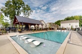 Located at 3110 Doe River Way in Matthews, NC, this outdoor amenity area is designed as a resort-style retreat for residents of the surrounding community. At its centerpiece is a generously sized swimming pool with crystal-clear turquoise water, offering ample space for both leisure and recreation. A distinctive shallow wading shelf lines one side of the pool, fitted with sculpted white in-water lounge chairs — ideal for relaxing while staying cool. A timber-framed covered pavilion with a standing-seam metal roof anchors one end of the pool deck, providing shaded gathering space with a stone accent wall and pendant lighting, adding a warm, rustic character to the overall design. The surrounding pool deck is finished with clean concrete pavers and outfitted with a generous collection of chaise lounges and umbrella-shaded seating areas, accommodating a variety of outdoor social activities. Black iron fencing encloses the entire area, providing a secure and well-defined boundary while maintaining open sightlines to the surrounding residential buildings and mature tree canopy. A separate fenced zone adjacent to the pool area appears to offer additional outdoor amenities, including a playground and dining-height patio furniture. The overall aesthetic blends craftsman and modern design elements cohesively.