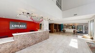 Located at 1223 N Haven Dr in Fayetteville, Arkansas, this single-floor community clubhouse offers a generous and thoughtfully designed gathering space. The entrance greets visitors with a prominent front desk clad in stacked brick, set against a bold red accent wall beneath soaring double-height ceilings adorned with a striking multi-arm chandelier. Light-toned tile flooring runs throughout the main corridor and lobby, creating a clean, open aesthetic.

The space transitions seamlessly into a sun-drenched lounge area featuring floor-to-ceiling windows and sliding glass doors that invite abundant natural light and views of the surrounding greenery. Vivid teal and green accent walls provide colorful focal points throughout, while a lush vertical garden panel adds a natural, modern touch near the entry hallway.

A large, open recreation area accommodates a billiard table, communal seating, and a bar-height counter beneath a "Stay Awhile" wall mural, fostering a social atmosphere. A cozy, sunken lounge nook features a bold botanical-print feature wall and deep, comfortable sectional seating, ideal for relaxed gatherings. The space also includes a well-appointed kitchenette and a staircase leading to an upper-level overlook. Together, these areas create a dynamic, welcoming environment designed for community connection and leisure.