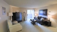 This single-floor space offers a comfortable and thoughtfully arranged living environment. The living room serves as the centerpiece, featuring plush wall-to-wall carpeting that adds warmth underfoot. A large sliding glass door floods the room with natural light and provides direct access to an outdoor area, creating a seamless indoor-outdoor connection. The textured ceiling adds a classic architectural touch, while neutral-toned walls provide a versatile backdrop for a variety of décor styles. The living area is generously proportioned, offering ample room for multiple seating arrangements. A bold, dark cabinet unit anchors one side of the room, providing functional storage while adding a striking contrast to the light-colored walls and carpet. Tasteful decorative accents, including framed wall art and a tall decorative vase, contribute to the polished, inviting ambiance. The layout is open and airy, allowing natural light to reach deep into the space. Overall, the space combines practical design with comfortable living, making it a welcoming environment well-suited for both relaxing and entertaining.
