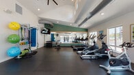 Located at 16920 SE Sunnyside Rd in Happy Valley, Oregon, this fitness center is a thoughtfully designed, single-floor exercise facility built to support a wide range of workout routines. The space features soaring vaulted ceilings with exposed industrial-style ductwork, lending the room an open, airy feel. Large windows along multiple walls allow generous amounts of natural light to fill the space, with views of an outdoor pool area and surrounding greenery visible from select vantage points. The flooring throughout is dark rubber matting, providing durability and comfort underfoot. A full-length mirror framed with a warm wood accent and set against a bold green accent wall reflects the cardio zone, which is equipped with multiple treadmills, elliptical machines, and a stationary bike. A cable resistance machine anchors a dedicated strength training area, complemented by a dumbbell rack and a functional fitness station outfitted with resistance bands, stability balls, exercise mats, and battle ropes. Wall-mounted televisions are positioned throughout for entertainment during workouts. Recessed lighting and ceiling fans contribute to a comfortable training environment. The overall ambiance is clean, modern, and energizing, making this facility a comprehensive fitness resource.