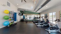 Located at 16920 SE Sunnyside Rd in Happy Valley, Oregon, this fitness center is a thoughtfully designed, single-floor exercise facility built to support a wide range of workout routines. The space features soaring vaulted ceilings with exposed industrial-style ductwork, lending the room an open, airy feel. Large windows along multiple walls allow generous amounts of natural light to fill the space, with views of an outdoor pool area and surrounding greenery visible from select vantage points. The flooring throughout is dark rubber matting, providing durability and comfort underfoot. A full-length mirror framed with a warm wood accent and set against a bold green accent wall reflects the cardio zone, which is equipped with multiple treadmills, elliptical machines, and a stationary bike. A cable resistance machine anchors a dedicated strength training area, complemented by a dumbbell rack and a functional fitness station outfitted with resistance bands, stability balls, exercise mats, and battle ropes. Wall-mounted televisions are positioned throughout for entertainment during workouts. Recessed lighting and ceiling fans contribute to a comfortable training environment. The overall ambiance is clean, modern, and energizing, making this facility a comprehensive fitness resource.