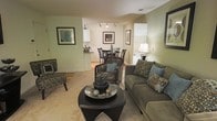 Located at 1140 Trinity Pine Ln in Raleigh, NC, this single-floor residence offers a thoughtfully designed layout that balances comfort and functionality. The living area serves as the heart of the home, featuring soft, neutral wall tones in warm sage and cream that create a calm and welcoming ambiance. Plush carpeting underfoot adds a cozy feel, while track lighting on the ceiling provides focused illumination throughout the space. The living room flows naturally into the dining area, where a classic round table and chair set sit beneath recessed lighting, making it an ideal spot for meals and gatherings. Just beyond, the kitchen is visible through an open pass-through, showcasing white cabinetry and light countertops that keep the space feeling bright and airy. The open-concept design ensures a seamless connection between the living, dining, and kitchen areas, enhancing the sense of spaciousness throughout the single floor. Framed wall art and carefully chosen furnishings add character and a polished, lived-in aesthetic, making this Raleigh residence a comfortable and inviting place to call home.