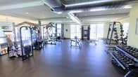 This fitness center offers a spacious, single-floor layout designed to accommodate a comprehensive workout experience. The room is outfitted with a variety of strength training machines, cable systems, and weight benches, thoughtfully arranged to allow ample room for movement between stations. The flooring consists of durable dark rubber matting, providing traction and cushioning suited for high-impact activities. Overhead, exposed industrial-style ductwork runs across the ceiling, lending the space an open, functional aesthetic while recessed lighting ensures consistent illumination throughout. Large windows and sliding glass doors line one wall, inviting abundant natural light and offering views of the surrounding outdoor environment. Full-length mirrors are positioned along an adjacent wall, a practical feature that allows users to monitor their form during exercise. The high ceiling height enhances the sense of openness, preventing any feeling of confinement. The overall ambiance is clean, bright, and purposeful, creating an environment well-suited to focused physical training. The combination of natural light, industrial design elements, and a well-curated equipment selection makes this fitness center a functional and inviting space for exercise.