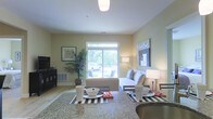 This single-floor residence showcases an open-concept design that seamlessly connects the dining and living areas, creating a cohesive and functional layout. The living room is anchored by a comfortable sofa dressed with decorative pillows, positioned to take full advantage of a large sliding window that fills the space with natural light and frames views of lush greenery outside. Light-toned walls and light hardwood-style flooring contribute to the bright, airy atmosphere throughout.

The dining area, visible in the foreground, features a dark-toned table set atop a bold black-and-white striped area rug, providing a striking contrast against the warmer tones of the room. A granite or stone-finish countertop edges the dining space, suggesting proximity to the kitchen. Accent lighting, including a ceiling-mounted fixture and a pair of table lamps, ensures the space remains warmly lit at all times.

Decorative elements such as framed wall art, a large potted palm plant, and carefully curated accessories add personality and warmth to the interior. The overall ambiance is clean, contemporary, and inviting, designed for comfortable everyday living.