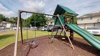 This single-story residential community offers a welcoming outdoor environment designed with comfort and recreation in mind. The standout feature of the property's exterior is a dedicated playground area, complete with a multi-function play structure finished in green and tan tones. The set includes a covered platform with a green canopy roof, an attached slide, climbing elements, and a separate swing set with two belt swings — all resting on a cushioned mulch surface for safety.

Surrounding the playground, well-maintained grassy lawns stretch across the grounds, framed by mature deciduous trees that provide generous shade and a sense of seclusion. The open-sky setting allows ample natural light to fill the outdoor spaces throughout the day. Nearby, a paved parking area is conveniently accessible, and the neighboring residential buildings are visible in the background, reflecting a traditional architectural style with neutral exteriors and balconies.

The overall ambiance of the community is friendly and family-oriented, with the outdoor amenities thoughtfully positioned to encourage active play and community engagement. The combination of green open space, established trees, and recreational features makes this property an appealing setting for residents of all ages.
