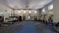 This generously sized fitness center offers a comprehensive workout environment designed for versatility and comfort. The space is defined by its soaring ceilings, which lend an open, airy atmosphere and are complemented by a large, industrial-style ceiling fan that ensures consistent air circulation throughout. Rows of clerestory windows line the upper walls, flooding the interior with natural light while maintaining privacy at floor level.

The main workout floor is covered in durable, patterned carpet tiles arranged in a herringbone layout, providing both cushioning and traction. A designated stretching and yoga area features a large blue mat at the center of the open floor space. Along one side, a row of treadmills and stationary bikes is positioned near wall-mounted flat-screen televisions, creating an engaging cardio zone. The opposite side houses a variety of strength training machines and free weight equipment, including cable machines, weight benches, and medicine balls.

Full-length mirrors are strategically placed across multiple walls, visually expanding the space and allowing users to monitor their form. An interior staircase with metal railings leads to an elevated mezzanine level, offering additional sightlines across the facility. Recessed lighting supplements the natural light, ensuring the space remains bright and functional at all hours.