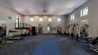 This generously sized fitness center offers a comprehensive workout environment designed for versatility and comfort. The space is defined by its soaring ceilings, which lend an open, airy atmosphere and are complemented by a large, industrial-style ceiling fan that ensures consistent air circulation throughout. Rows of clerestory windows line the upper walls, flooding the interior with natural light while maintaining privacy at floor level.

The main workout floor is covered in durable, patterned carpet tiles arranged in a herringbone layout, providing both cushioning and traction. A designated stretching and yoga area features a large blue mat at the center of the open floor space. Along one side, a row of treadmills and stationary bikes is positioned near wall-mounted flat-screen televisions, creating an engaging cardio zone. The opposite side houses a variety of strength training machines and free weight equipment, including cable machines, weight benches, and medicine balls.

Full-length mirrors are strategically placed across multiple walls, visually expanding the space and allowing users to monitor their form. An interior staircase with metal railings leads to an elevated mezzanine level, offering additional sightlines across the facility. Recessed lighting supplements the natural light, ensuring the space remains bright and functional at all hours.