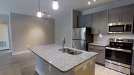 This single-floor space showcases a thoughtfully designed open-concept kitchen and living area. The kitchen serves as the focal point, anchored by a spacious island with speckled granite countertops and a double stainless steel sink. Pendant lights hang above the island, complementing a track lighting system that provides layered illumination throughout the space. Grey shaker-style cabinetry lines the walls, offering ample storage while contributing to the contemporary aesthetic. A full-sized stainless steel refrigerator and white subway tile backsplash add polished, modern touches. The warm-toned hardwood flooring flows seamlessly from the kitchen into the adjoining living area, enhancing the open feel of the layout. Neutral wall tones throughout create a versatile backdrop that complements the space's modern finishes. The open layout between the kitchen and living area promotes a connected, functional flow ideal for both everyday living and entertaining. Overall, the space conveys a clean, contemporary design characterized by quality materials and a cohesive color palette of greys, whites, and warm wood tones.