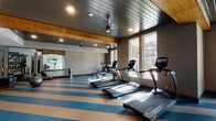 This fitness center offers a comprehensive and thoughtfully designed workout environment across a single open floor. The space is immediately distinguished by its bold blue and tan striped flooring, which adds a dynamic visual energy while providing a durable, easy-to-maintain surface throughout the facility.

The cardio zone is lined with True-brand treadmills and elliptical machines, each equipped with individual screens for entertainment during workouts. Large windows along the walls flood the space with natural light and offer views of the surrounding exterior, creating an open, airy atmosphere. A full-length mirror along one wall further amplifies the sense of space.

A dedicated functional training area houses an extensive rack system stocked with medicine balls, kettlebells, and battle ropes, along with TRX suspension trainers and stability balls, supporting a wide range of strength and conditioning exercises. A wall-mounted flat-screen TV provides additional entertainment options in the open activity area.

The ceiling features a striking combination of dark metal paneling and warm wooden beam accents, lending the space a modern yet welcoming character. Ceiling fans and recessed lighting ensure comfortable conditions at all times. A water fountain and waste receptacle are conveniently positioned near the interior corridor, which also provides access to an additional private room.