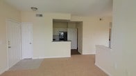 This single-floor residence offers a clean and functional layout designed for comfortable everyday living. The main living area features wall-to-wall carpeting in a neutral tone, creating a warm and inviting atmosphere. Soft, light-colored walls run throughout, contributing to a bright and airy feel.

The kitchen is positioned at the heart of the space, partially enclosed by a raised breakfast bar with dark countertops that provides a natural dividing point between the kitchen and the living area. White cabinetry lines the upper walls of the kitchen, offering ample storage, while black appliances — including a refrigerator and what appears to be a range — add a modern contrast. The kitchen flooring transitions to a darker material, visually distinguishing it from the carpeted living space.

A hallway visible beyond the kitchen suggests additional rooms or private spaces deeper within the unit. The layout flows naturally from the main living area into the kitchen and beyond. Overhead lighting and a ceiling vent indicate a central climate control system. Overall, the space presents a straightforward and practical design with a cohesive neutral color palette throughout.