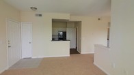 This single-floor residence offers a clean and functional layout designed for comfortable everyday living. The main living area features wall-to-wall carpeting in a neutral tone, creating a warm and inviting atmosphere. Soft, light-colored walls run throughout, contributing to a bright and airy feel.

The kitchen is positioned at the heart of the space, partially enclosed by a raised breakfast bar with dark countertops that provides a natural dividing point between the kitchen and the living area. White cabinetry lines the upper walls of the kitchen, offering ample storage, while black appliances — including a refrigerator and what appears to be a range — add a modern contrast. The kitchen flooring transitions to a darker material, visually distinguishing it from the carpeted living space.

A hallway visible beyond the kitchen suggests additional rooms or private spaces deeper within the unit. The layout flows naturally from the main living area into the kitchen and beyond. Overhead lighting and a ceiling vent indicate a central climate control system. Overall, the space presents a straightforward and practical design with a cohesive neutral color palette throughout.