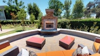 This inviting outdoor communal space is thoughtfully designed to accommodate a variety of social and leisure activities. The area is organized into several distinct zones, each serving a specific purpose while contributing to the overall cohesive ambiance.

At the heart of the space is a covered lounge area furnished with wicker seating accented by vibrant orange cushions, providing a comfortable retreat from the sun. Adjacent to this is a dedicated dining zone featuring a wicker table with matching chairs, shaded by a large overhead umbrella — perfect for al fresco meals.

A standout feature of the space is the stone-built outdoor fireplace, set within a semi-circular seating enclosure with low perimeter walls, creating an intimate gathering spot ideal for cooler evenings. Nearby, a fully equipped outdoor kitchen includes a built-in BBQ grill and counter space, complemented by a high-top dining table with bar-height seating for casual outdoor dining.

The grounds are beautifully landscaped with manicured lawns, flowering trees, vibrant shrubbery, and mature trees that provide natural shade and privacy. Decorative lamp posts add a charming architectural touch throughout. The entire space benefits from abundant natural light, an open-sky atmosphere, and a warm, welcoming character suited for both relaxation and community gatherings.