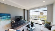 Located along the Grand River corridor near I-196 in Grand Rapids, Michigan, this single-floor unit offers a thoughtfully designed contemporary interior with striking panoramic views of the river and surrounding cityscape. The living area is defined by expansive floor-to-ceiling windows with black steel framing, flooding the space with natural light while providing unobstructed sightlines to the water and downtown beyond. Hardwood-style flooring runs throughout, complementing the clean, neutral color palette of gray and white tones.

The kitchen is well-appointed with light gray cabinetry extending to the ceiling, stainless steel appliances including a range, microwave, and refrigerator, and a sleek white quartz peninsula with bar seating. A dark tile backsplash and pendant lighting add visual depth, while a large abstract mural anchors the adjacent dining area with a bold artistic statement.

The bedroom is generously proportioned, featuring a large window that continues the river view theme, creating a serene, light-filled retreat. The bathroom offers a clean, minimalist design with white cabinetry and warm wood-tone flooring. Additional features include in-unit laundry and ample storage, making this an efficiently organized and visually refined urban residence.