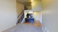 This two-story residence is designed with an open, airy layout that maximizes the sense of space across both floors. The main level features a seamless transition between the living area and kitchen, highlighted by soaring double-height ceilings that create a dramatic vertical openness rarely found in comparable homes. Plush carpeting anchors the living space, while the kitchen and surrounding areas feature warm hardwood-style flooring, providing a clear and stylish distinction between zones. The kitchen is equipped with dark cabinetry, black appliances, and a peninsula breakfast bar with a bold blue base — a standout design element that adds character to the space. A staircase with sleek metal railings leads to the upper level, which overlooks the main living area through an open balcony railing, maintaining the sense of connectivity between floors. Neutral, warm-toned walls throughout contribute to a cohesive and welcoming ambiance. Multiple interior doors on the main level suggest well-defined secondary spaces, adding to the functional versatility of the floor plan. The combination of elevated ceilings, open-concept design, and thoughtful material choices makes this a visually impressive and highly livable two-floor space.
