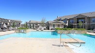 Located at 6610 34th St in Lubbock, Texas, this outdoor amenity space delivers a resort-like experience designed for both relaxation and socializing. The centerpiece is a generously sized swimming pool with clear, blue water, accented by a decorative water feature that adds a tranquil ambiance to the surroundings. The pool deck is finished in natural stone tile, providing a durable and visually appealing surface that complements the overall aesthetic. Poolside lounge chairs are thoughtfully positioned to take full advantage of the West Texas sunshine and open sky views.

Adjacent to the pool, a beautifully crafted pergola structure offers a shaded outdoor gathering area. Built with stacked stone columns and topped with a slatted wood canopy, the pergola shelters an outdoor kitchen and bar counter finished with granite-style surfaces — ideal for entertaining. Comfortable seating is arranged beneath the structure, creating an inviting space for residents to unwind.

The amenity area is enclosed by a classic black wrought iron fence, beyond which a manicured green lawn stretches openly, framed by multi-story residential buildings with brick and stone exteriors. Gooseneck-style outdoor lighting adds a refined, community-oriented character to the space, completing a well-appointed outdoor retreat.