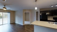 This single-floor residence offers a thoughtfully designed open-concept layout that seamlessly connects the kitchen and living areas. The kitchen is anchored by a granite-topped breakfast bar with a pendant light overhead, providing both functional counter space and a casual dining option. Dark hardwood flooring runs throughout the main living area, creating a rich contrast against the neutral beige walls and crisp white trim. A built-in open shelving pantry is conveniently positioned near the kitchen, offering practical storage within easy reach. The transition from the living area leads into a carpeted bedroom, visible through a doorway and featuring a ceiling fan, adding a soft and comfortable feel to the private sleeping space. The layout is straightforward and efficient, with clean sightlines from the kitchen across the main living space. Recessed HVAC venting in the ceiling contributes to a clean, uncluttered aesthetic overhead. The overall ambiance is calm and neutral, with an understated palette that provides a versatile backdrop well-suited to a variety of interior styles and furnishing choices.