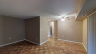 This single-floor space presents a clean, straightforward layout defined by its distinctive parquet hardwood flooring, which adds warmth and visual texture throughout the open living area. The flooring's geometric pattern creates a classic aesthetic that anchors the neutral color palette of the walls, which transition between a cool gray and a warm beige tone, lending the space a subtle sense of depth and definition.

The main living area is open and uncluttered, providing a versatile canvas for various furniture arrangements. A ceiling fan with an integrated light fixture is centrally mounted, offering both ambient lighting and air circulation. White baseboards run consistently along the walls, providing clean contrast against the flooring and wall tones.

A partial wall divides the main living space, creating a natural transition toward a hallway that leads further into the unit. At the end of the hallway, built-in shelving is visible, hinting at additional storage solutions within the space. The overall ambiance is calm and neutral, making it a practical and adaptable environment suitable for a range of uses.