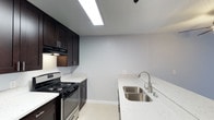 This well-appointed single-floor unit showcases a cohesive modern design throughout its living spaces. The kitchen is a standout feature, outfitted with rich dark espresso cabinetry that extends floor to ceiling, offering generous storage. Gleaming quartz countertops with subtle veining provide ample prep space, while a stainless steel gas range and a deep double-basin sink round out the functional layout. A breakfast bar with seating overhang connects the kitchen seamlessly to the living area, which is finished with wide-plank dark hardwood flooring that flows throughout. The living space is brightened by natural light and features a ceiling fan overhead, creating a comfortable and airy atmosphere. The bedroom areas are similarly finished with the same rich hardwood floors, neutral light-gray walls, and textured ceilings, providing a clean and versatile backdrop. A walk-in closet offers practical storage. The bathroom is neatly finished with a full tub-shower combo, a dark-vanity with quartz countertop, and light tile flooring, maintaining the consistent design language found throughout the unit. Clean white baseboards and soft neutral tones unify each room, resulting in a polished and move-in-ready living environment.