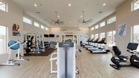 This fitness center at Christopher Todd Communities is a thoughtfully designed, single-floor facility built to support a comprehensive workout experience. The main gym area boasts soaring ceilings lined with a row of clerestory windows that flood the space with abundant natural light, while large ceiling fans maintain comfortable airflow throughout. Light wood-style flooring runs across the open floor plan, giving the space a clean, modern aesthetic.

The main workout floor is generously equipped with Life Fitness strength machines, free weights, benches, and a cable machine system, with full-length mirrors along one wall to support proper form. A dedicated cardio zone features multiple treadmills and additional cardio equipment positioned in front of large windows overlooking the community's outdoor pool area and palm-lined surroundings — providing an energizing view during workouts.

A separate cycling studio houses a row of spin bikes facing a mirrored wall, accented by a bold blue accent wall bearing the community's tagline, "A New Way To Live." Recessed lighting keeps this room well-lit for focused group or individual sessions.

The facility is accessed through large glass-panel double doors, reinforcing the bright and welcoming atmosphere. Overall, the space combines functionality with a polished, community-oriented design.