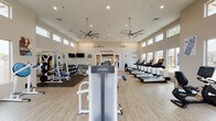 This fitness center at Christopher Todd Communities is a thoughtfully designed, single-floor facility built to support a comprehensive workout experience. The main gym area boasts soaring ceilings lined with a row of clerestory windows that flood the space with abundant natural light, while large ceiling fans maintain comfortable airflow throughout. Light wood-style flooring runs across the open floor plan, giving the space a clean, modern aesthetic.

The main workout floor is generously equipped with Life Fitness strength machines, free weights, benches, and a cable machine system, with full-length mirrors along one wall to support proper form. A dedicated cardio zone features multiple treadmills and additional cardio equipment positioned in front of large windows overlooking the community's outdoor pool area and palm-lined surroundings — providing an energizing view during workouts.

A separate cycling studio houses a row of spin bikes facing a mirrored wall, accented by a bold blue accent wall bearing the community's tagline, "A New Way To Live." Recessed lighting keeps this room well-lit for focused group or individual sessions.

The facility is accessed through large glass-panel double doors, reinforcing the bright and welcoming atmosphere. Overall, the space combines functionality with a polished, community-oriented design.