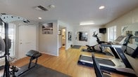 This dedicated fitness center offers a comprehensive workout environment spread across a single floor. The space features warm hardwood flooring throughout, complemented by neutral gray walls and a textured ceiling with recessed lighting and fluorescent fixtures that ensure bright, even illumination across the entire area.

The cardio zone is well-stocked with a treadmill, elliptical trainer, and a recumbent stationary bike, each positioned to take advantage of the natural light streaming in through multiple windows fitted with horizontal blinds. Views of mature trees and greenery outside add a refreshing outdoor connection during workouts.

The strength-training area houses a multi-station cable machine, a weight bench, and a kettlebell, catering to a full range of resistance exercises. Large wall-mounted mirrors, accented with motivational text and graphics, visually expand the space and allow users to monitor their form. A wall-mounted flat-screen television provides entertainment options during training sessions.

An adjacent area, visible through an open passageway, includes a small restroom or utility space, adding convenience to the facility. A water cooler is accessible nearby, and a fire extinguisher is mounted on the wall for safety. Protective rubber floor mats are placed beneath the equipment throughout. The overall ambiance is clean, functional, and conducive to focused, effective training sessions.