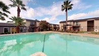 This amenity space features two distinct areas designed to support an active lifestyle. The outdoor pool area is a standout highlight, showcasing a generously sized swimming pool with clear turquoise water, surrounded by a spacious sun deck lined with lounge chairs. Mature palm trees and flowering crape myrtles frame the area, adding a lush, resort-like atmosphere beneath open skies. The multi-story residential buildings encircling the pool create a private, courtyard-style setting.

The fitness center is a well-appointed, single-floor gym space outfitted with rubber flooring throughout for safety and durability. It is divided into dedicated zones for cardiovascular and strength training. The cardio area features multiple treadmills and elliptical machines, with large windows offering views of the pool area and palm trees beyond, allowing natural light to fill the space. The strength training section is equipped with a dual cable crossover machine, a lat pulldown station, a flat bench, and additional weight machines, providing a comprehensive range of workout options. Wall-mounted exercise guides assist users with proper form. The fitness center benefits from both natural light and overhead lighting, creating a bright and functional environment for daily workouts.