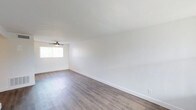 This single-floor space presents a clean, modern aesthetic defined by its crisp white walls and smooth ceilings that amplify the sense of openness throughout. The main living area is finished with light-toned hardwood-style flooring that runs seamlessly across the room, lending warmth and a contemporary feel underfoot. A ceiling fan is centrally positioned, providing both functional air circulation and a subtle design detail that complements the minimalist interior. Natural light filters in through a window fitted with vertical blinds, brightening the space and enhancing its airy, welcoming ambiance. White baseboards frame the perimeter of the room, adding a polished finishing touch that ties the interior together. The overall layout is open and unobstructed, offering flexible use of the space to suit a variety of needs. The neutral color palette and clean lines create a versatile backdrop that is well-suited to a range of interior styles and furnishings. The space presents itself in a well-maintained, move-in ready condition, making it an accessible and practical option for those seeking a straightforward, well-lit living environment.