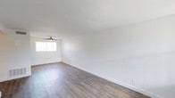 This single-floor space presents a clean, modern aesthetic defined by its crisp white walls and smooth ceilings that amplify the sense of openness throughout. The main living area is finished with light-toned hardwood-style flooring that runs seamlessly across the room, lending warmth and a contemporary feel underfoot. A ceiling fan is centrally positioned, providing both functional air circulation and a subtle design detail that complements the minimalist interior. Natural light filters in through a window fitted with vertical blinds, brightening the space and enhancing its airy, welcoming ambiance. White baseboards frame the perimeter of the room, adding a polished finishing touch that ties the interior together. The overall layout is open and unobstructed, offering flexible use of the space to suit a variety of needs. The neutral color palette and clean lines create a versatile backdrop that is well-suited to a range of interior styles and furnishings. The space presents itself in a well-maintained, move-in ready condition, making it an accessible and practical option for those seeking a straightforward, well-lit living environment.