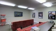 This single-floor space is designed as a casual lounge and media room, offering a welcoming and entertainment-focused environment. The room is finished with smooth, light-painted walls and features flush-mounted rectangular ceiling lights that provide even, bright illumination throughout the space.

At the heart of the room sits a large, L-shaped sectional sofa upholstered in a rich, textured burgundy fabric, complemented by small round side tables that add functional surface space. A wall-mounted flat-screen TV serves as the focal point on one wall, making the area well-suited for movie screenings, casual gatherings, or relaxed viewing sessions.

The walls are adorned with a curated selection of framed artwork and iconic movie posters, including recognizable classics such as Caddyshack and Star Wars, lending the space a pop-culture character and a sense of personality. A glass-top coffee table with a wire base sits at the center of the seating arrangement, adding a modern touch.

The flooring appears to be a neutral, smooth surface that ties the room together cohesively. The overall ambiance is casual yet thoughtfully arranged, making this space ideal for communal entertainment or social interaction in a comfortable, visually engaging setting.